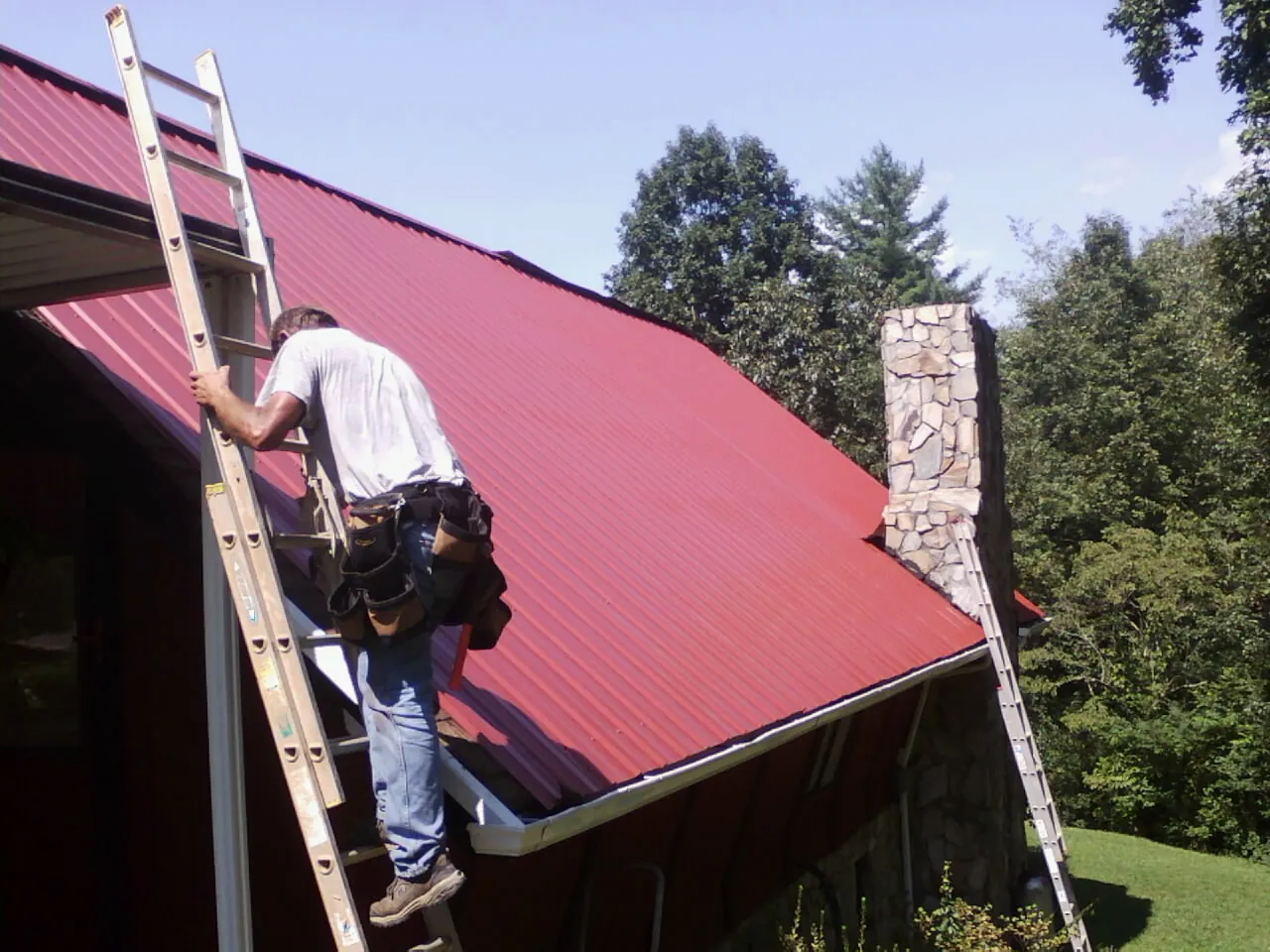 Roofer on a ladder beside a red metal roof and stone chimney — Soffit & Fascia Repair by Roofing Brothers in Los Angeles